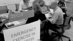 Crisis exercises- black and white photo of people gathered around a table under a sign that says 